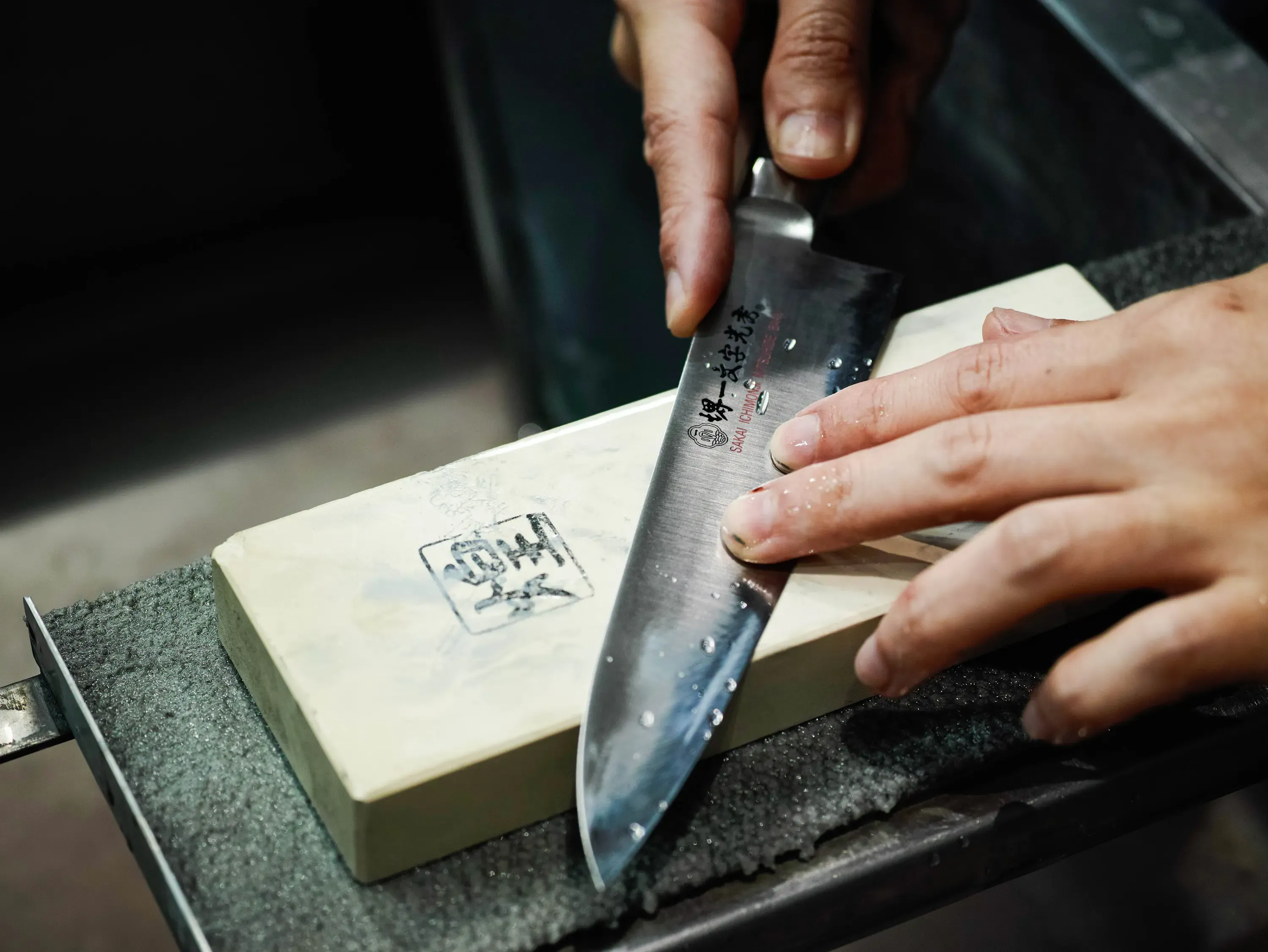 A maker's hands drawing a blade across a Japanese water stone, the edge held to an exact angle